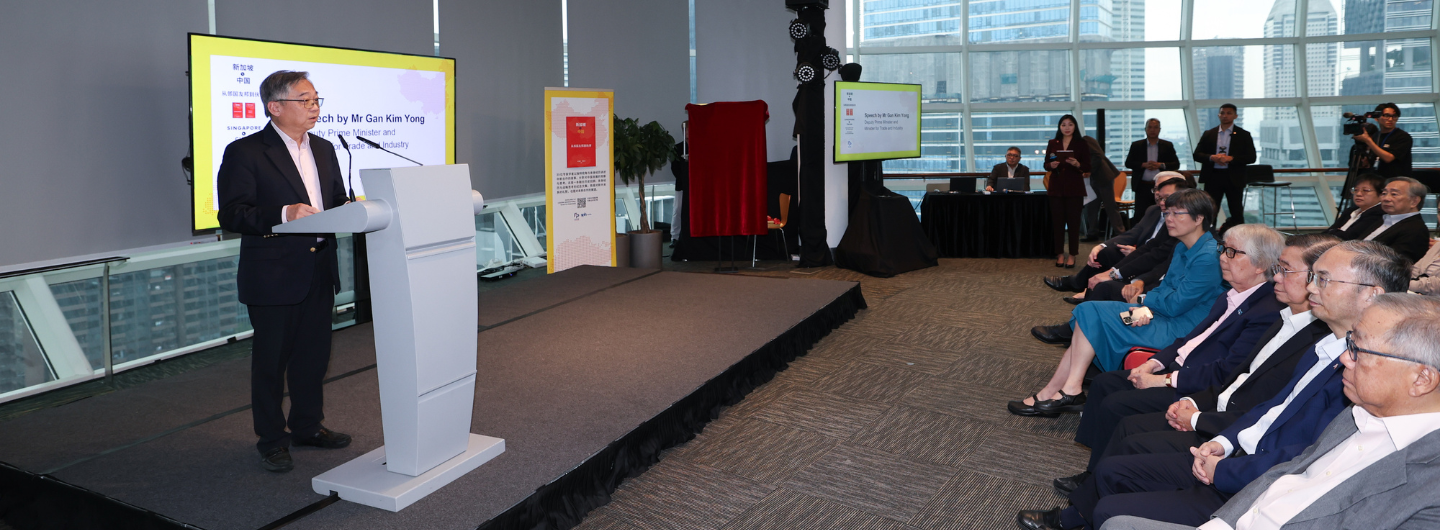 Speaker at podium addresses seated audience at indoor event; city skyline visible through large windows.